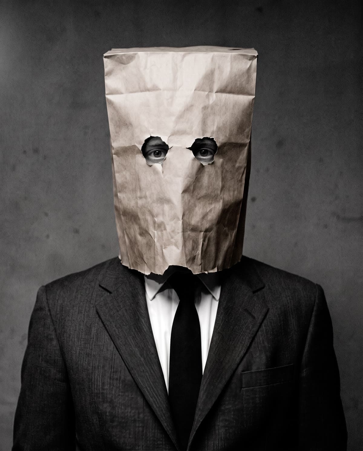 Retrato em preto e branco do personagem: terno escuro, gravata e uma sacola de papel cobrindo o rosto